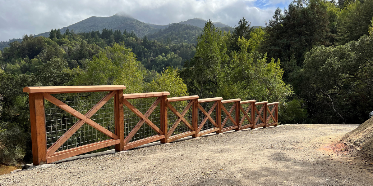 Fencing lines a dirt roadway overlooking misty, tree-covered mountains. 