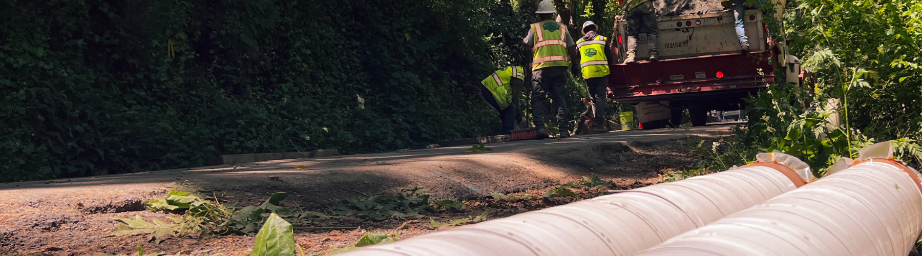 Two white pipes sit on the side of the road in the foreground. In the background, a construction crew works on a trench.