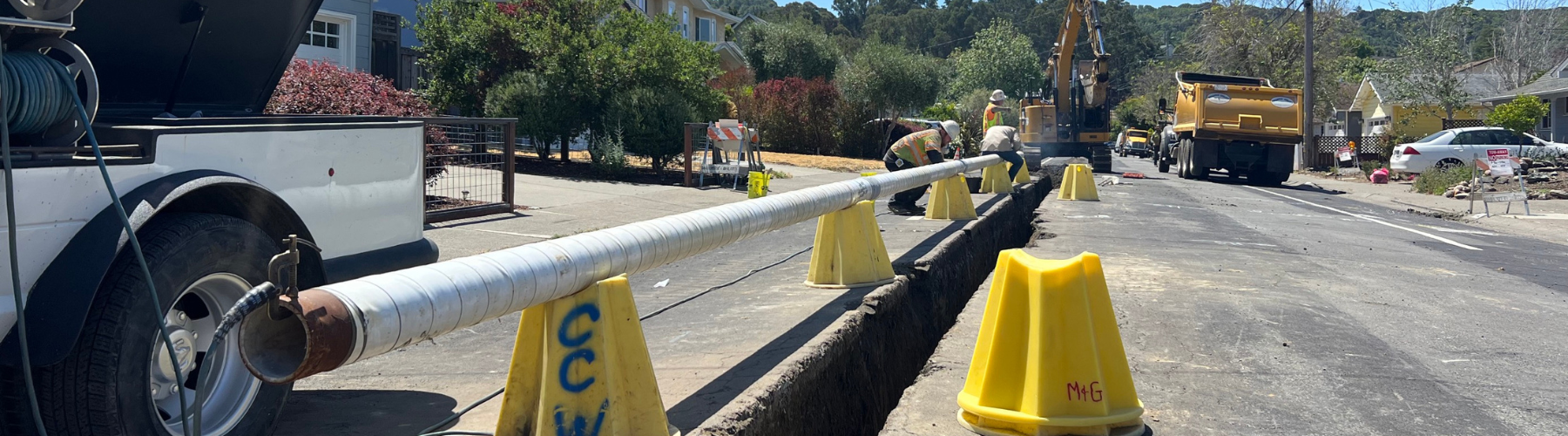 A water pipeline section sits on blocks above a trench in a residential neighborhood street.