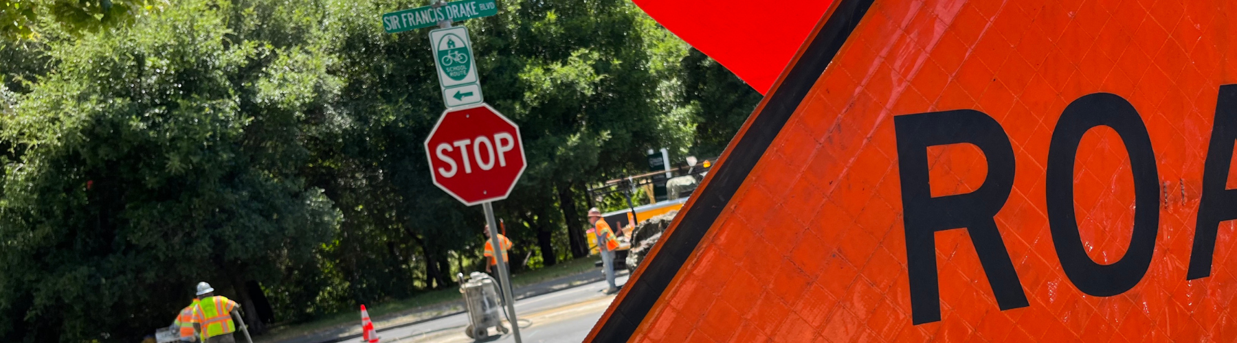 An orange road work sign in the foreground gives way to a view construction workers and traffic.