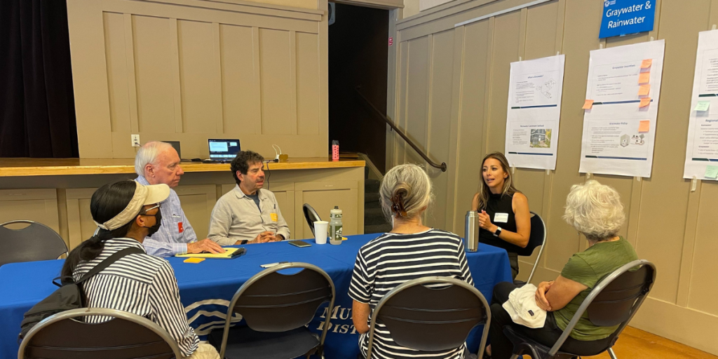 People sit around a table with a blue tablecloth. Posters with information appear on the wall behind.