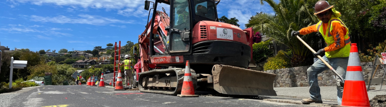 A person in a hard hat and bright clothing shovels dirt from a trench in a cone-marked construction zone nearby heavy equipment.