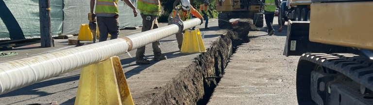 A water pipeline is prepared to be lowered into a trench in a roadway by construction workers.