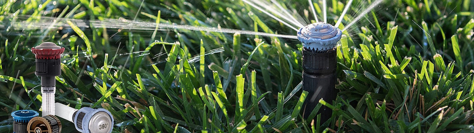 Image shows a group of sprinkler nozzles on the left, another working sprinkler on the left, all with a grassy backdrop.